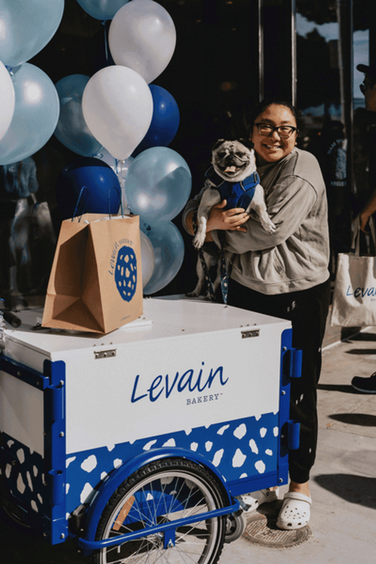 Woman holding a pug next to a Levain Bakery cookie cart with blue and white balloons, a branded bag, and an outdoor event setting.