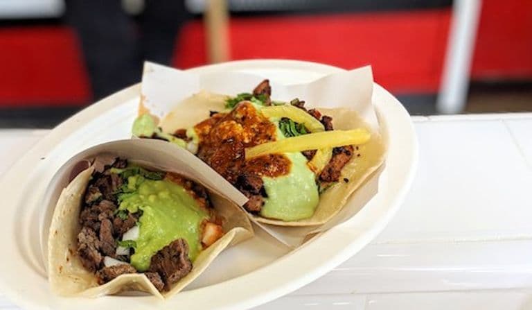 A close-up of two street-style tacos served on a white paper plate. One taco is filled with grilled meat, guacamole, and diced onions, while the other features grilled meat, guacamole, salsa, and a strip of pineapple, all on soft corn tortillas. The background shows a red and black counter, slightly out of focus.