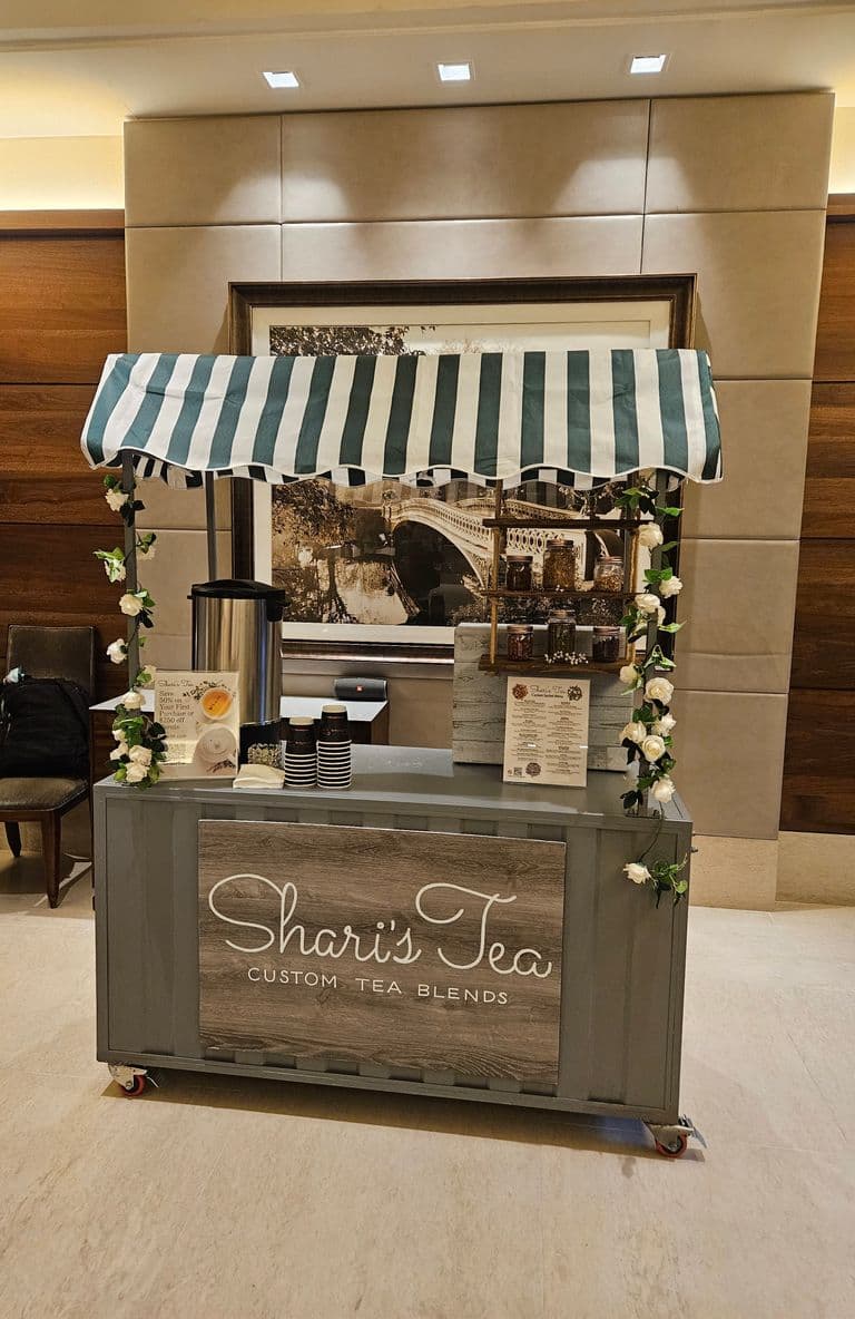 A charming mobile tea cart with a striped canopy and floral accents, featuring “Shari’s Tea – Custom Tea Blends,” offering hot tea service with jars of loose-leaf blends and takeaway cups.