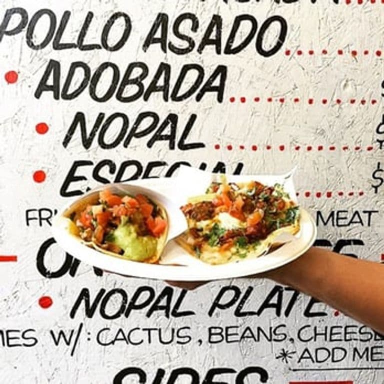 A person holds a white paper plate with two tacos topped with guacamole, diced tomatoes, onions, and cilantro in front of a textured wall menu. The menu lists items like "POLLO ASADO," "ADOBADA," "NOPAL," and "ESPECIAL" in bold black and red lettering.