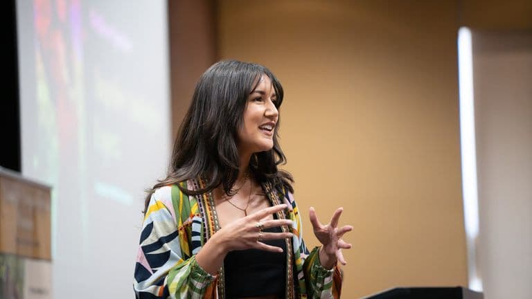 Woman speaking passionately at a podium in a colorful jacket, mid-gesture, with a blurred projection behind her.