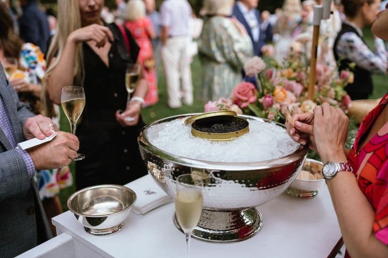 Close-up of a caviar bar setup with a large silver bowl filled with ice and an open tin of caviar, surrounded by guests holding champagne glasses at an elegant outdoor event.