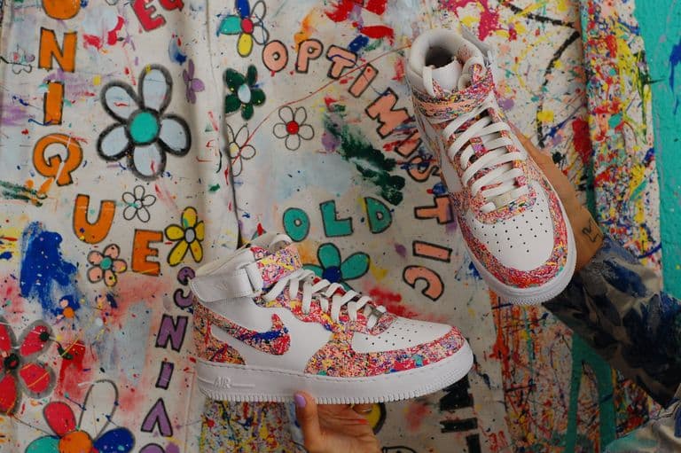 Two custom white sneakers with colorful splatter-paint designs are held up in front of a vibrant, graffiti-style backdrop featuring flowers and words like "unique" and "optimistic."