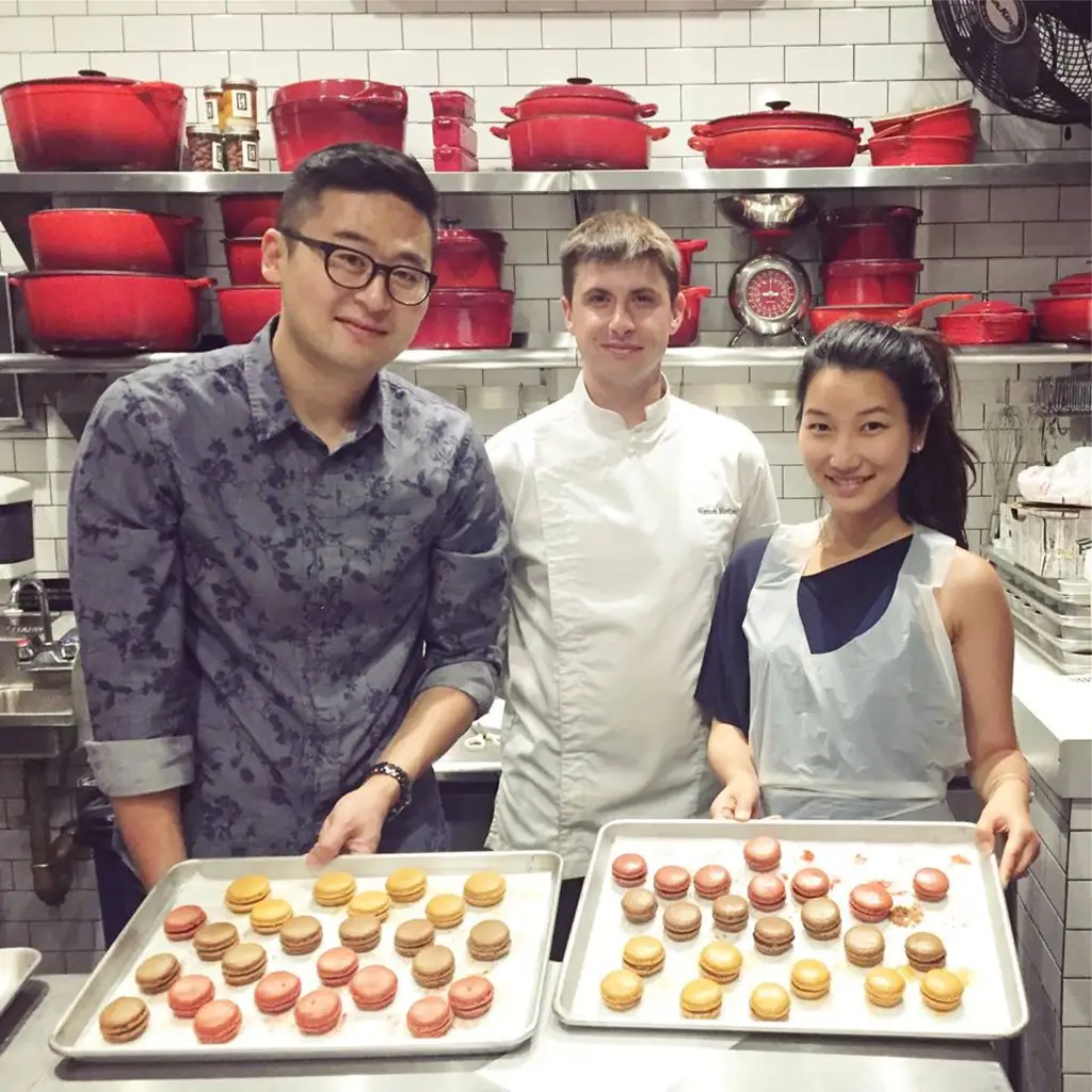 Two people holding trays of colorful macarons, posing with a chef in a kitchen.