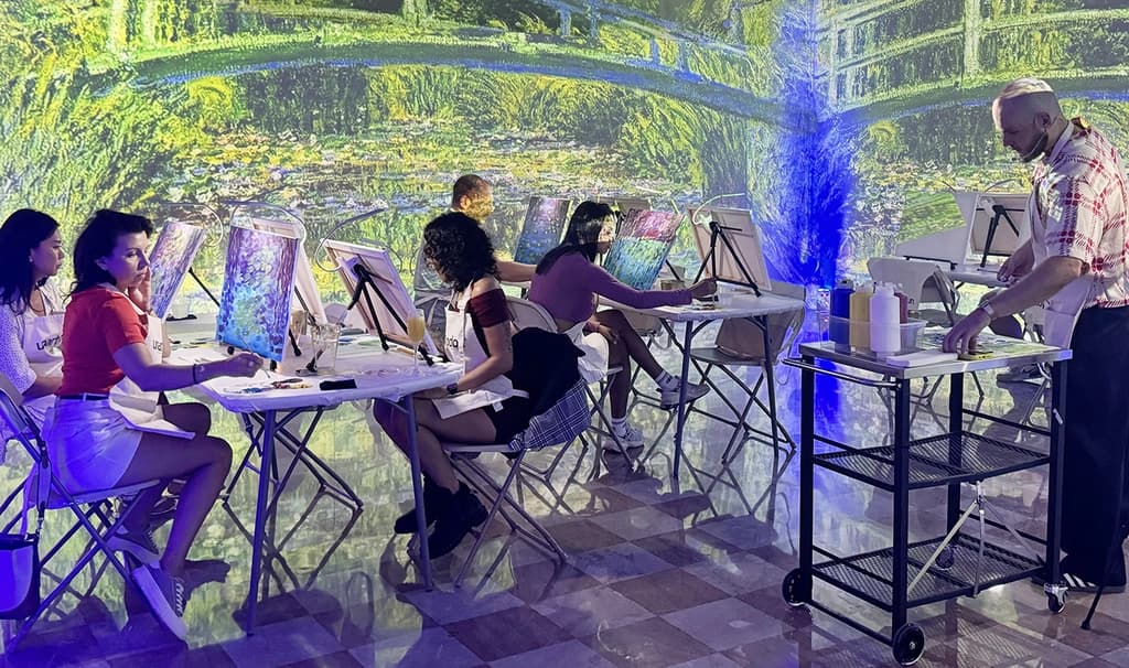 Two women seated at a table, painting a colorful Monet-style scene on their canvases, focused and enjoying the immersive environment.