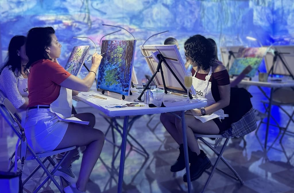 Participants at an art class, painting vibrant Monet-style artwork at easels, while a man in the background prepares painting supplies.