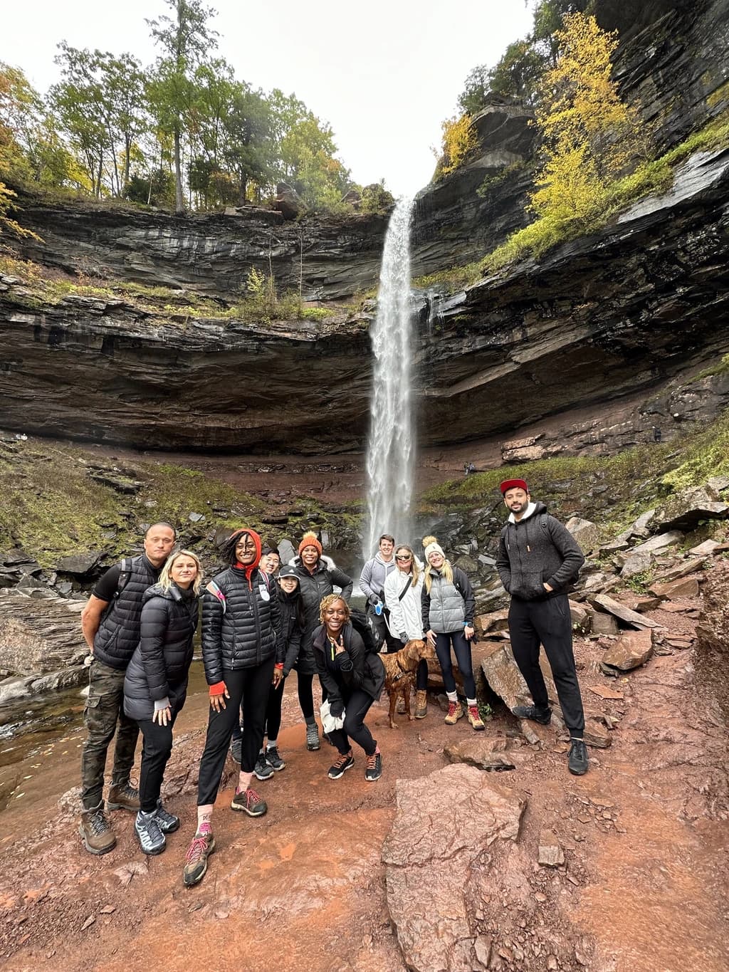 A group of hikers posing in front of a tall waterfall, surrounded by rocky terrain and fall foliage.