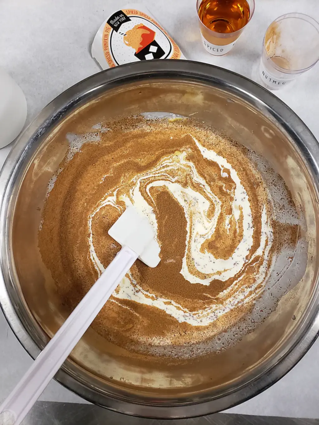 A large metal mixing bowl filled with spiced ice cream mixture, featuring ingredients like spiced whiskey and nutmeg nearby.