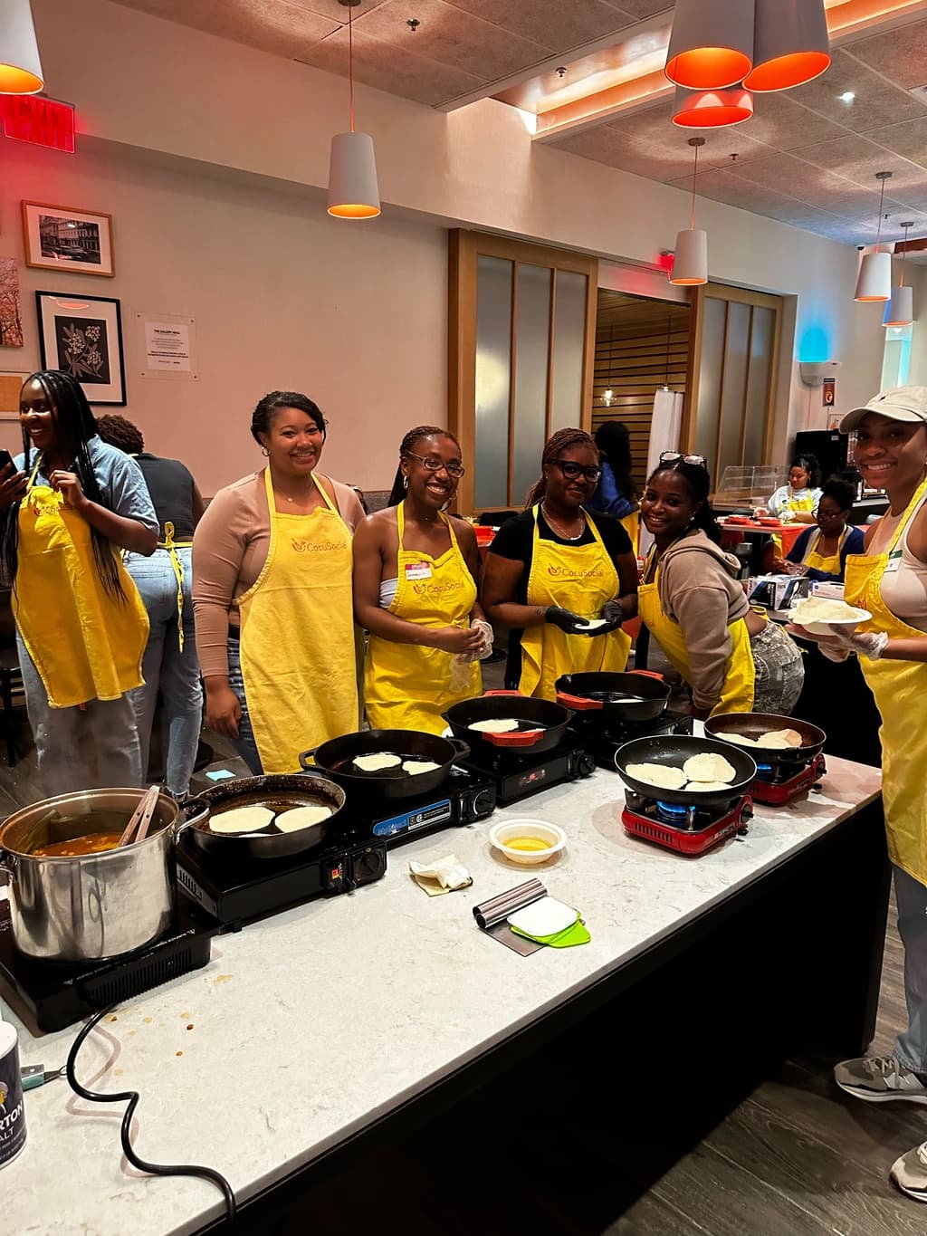 A group of smiling participants in yellow aprons standing around a cooking station, preparing food on portable stoves during a lively cooking class.
