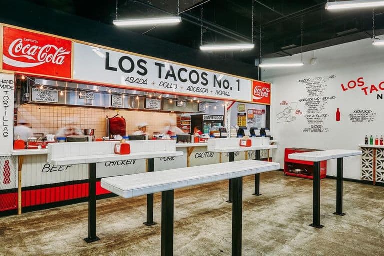 Interior of Los Tacos No. 1 with tiled standing tables, a visible open kitchen, and menu signage featuring taco options like beef, chicken, pork, and cactus.