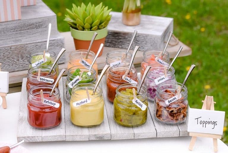 Mason jars filled with hot dog toppings like ketchup, mustard, relish, onions, bacon, chili, jalapeños, BBQ sauce, and more.