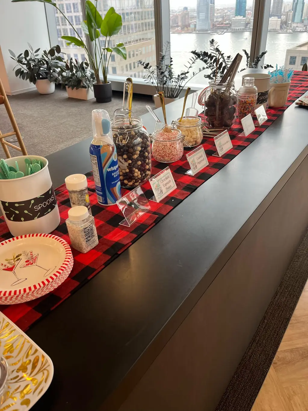 A toppings bar with jars of candies, whipped cream, and festive decor, set against a window view of a city and river.