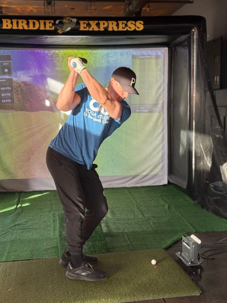 Person in mid-backswing stance at an indoor golf simulator, standing on artificial turf with a golf ball teed up in front of them, illuminated by natural light.