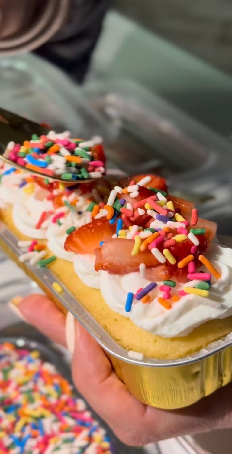 Vanilla loaf cake with whipped cream, fresh strawberries, and rainbow sprinkles being added at a dessert station.