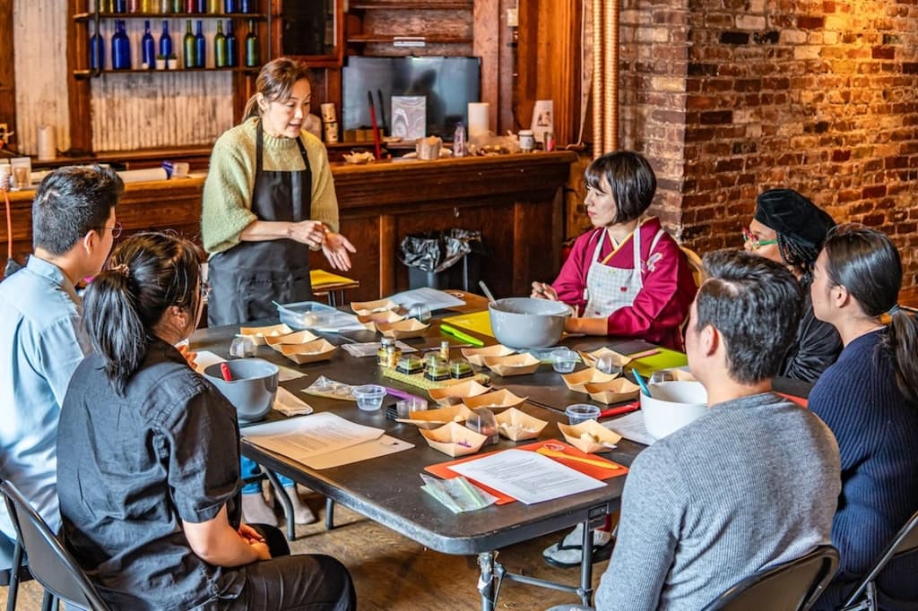 Group of people participating in a Japanese sweets making class, listening attentively to the instructor in a rustic setting.