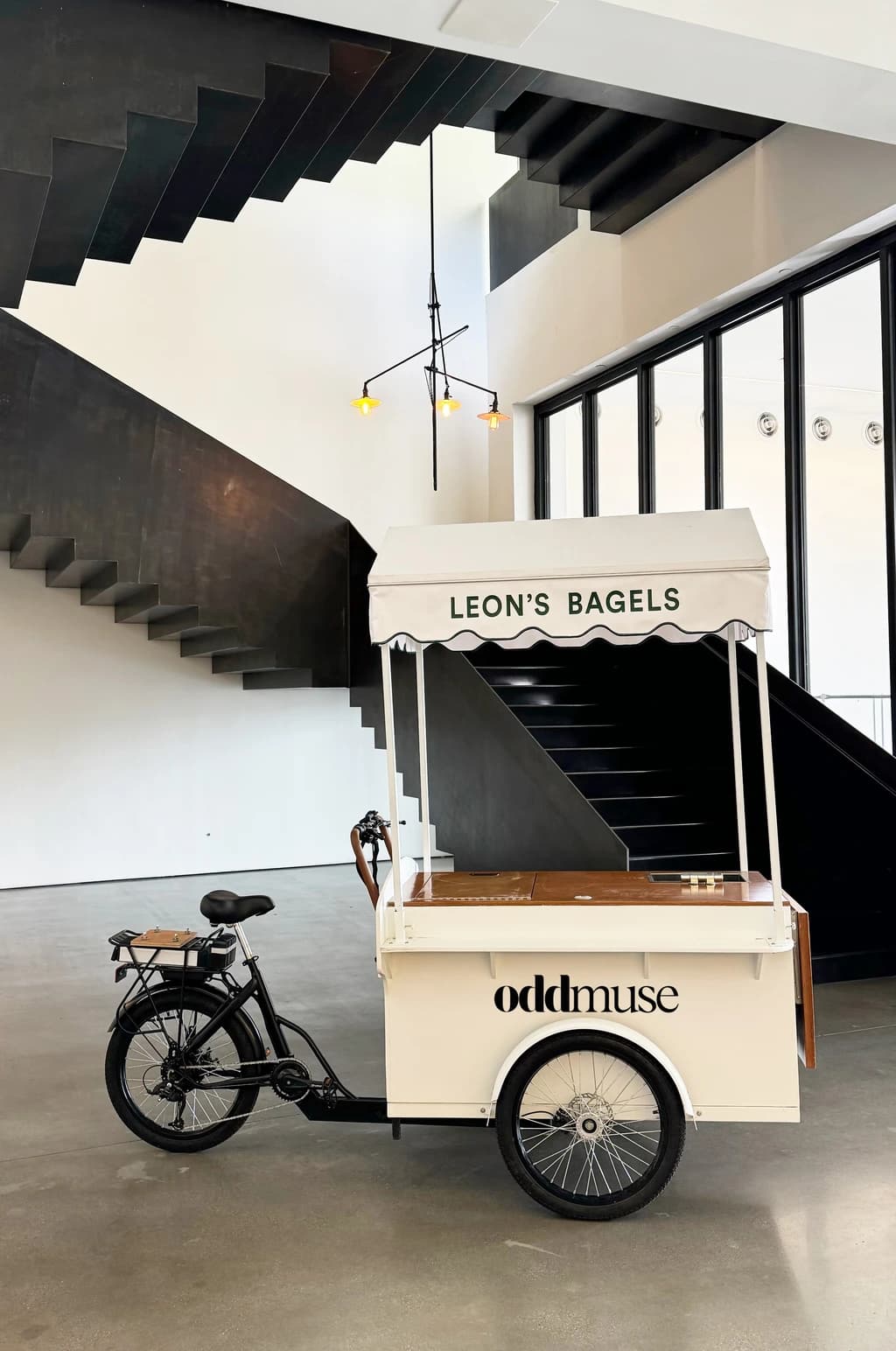 A 'Leon's Bagels' cart with a wooden countertop, parked outside a green garage door, framed by red brick walls and yellow bollards.
