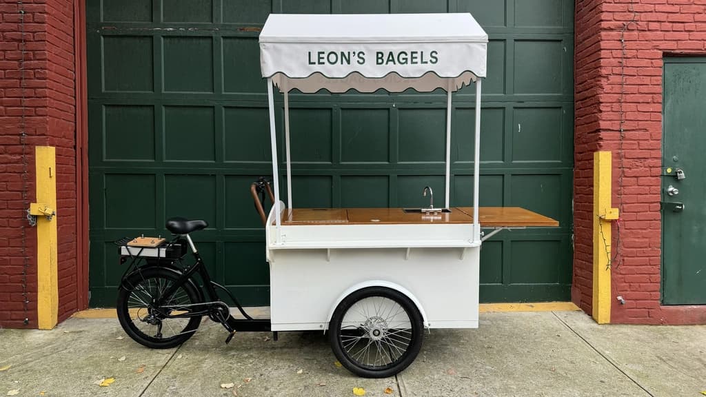 A sleek 'Leon's Bagels' cart with a canopy and bicycle, set in a modern space with black stairs and industrial lighting overhead.