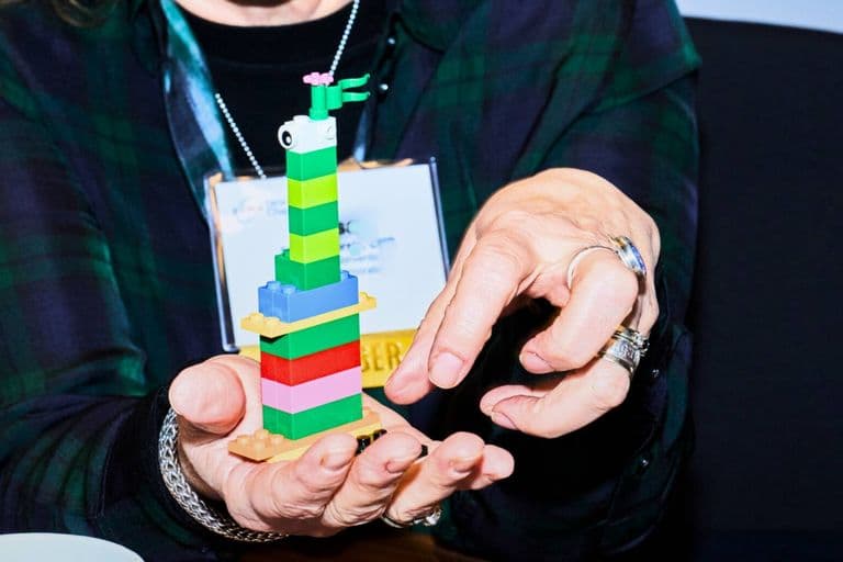 Close-up of a person holding a small, colorful LEGO tower decorated with flags and eyes, gesturing toward it with their other hand