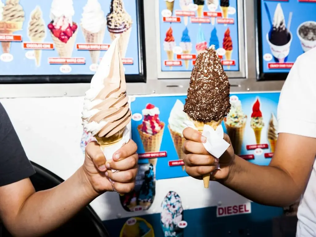 Two people holding ice cream cones from Mister Softee. One cone has a chocolate and vanilla swirl, and the other is chocolate-dipped with sprinkles. The background shows the ice cream truck's colorful menu.