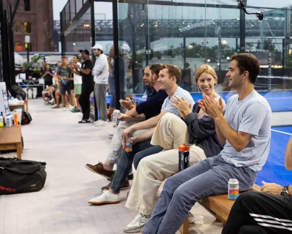Spectators sitting and clapping on the sidelines of a padel court, enjoying the game with smiles and refreshments.