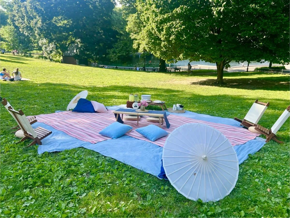 A picnic setup with a blue and pink striped mat, pillows, a small table with food, and parasols, set in a green park.