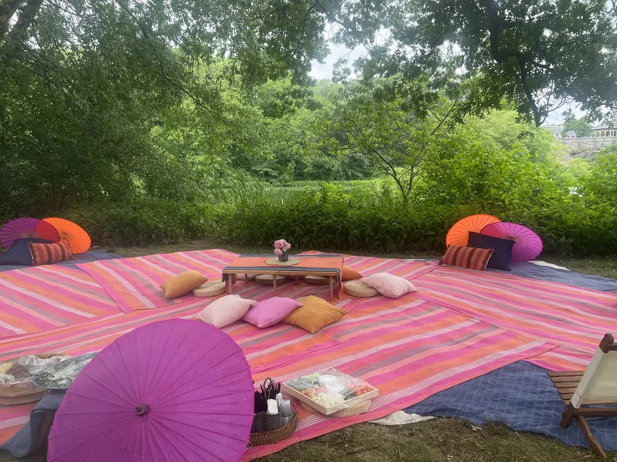 A picnic setup with colorful mats, pillows, and parasols under the shade of trees.