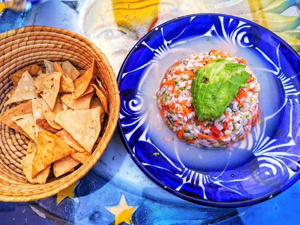 A colorful plate of ceviche topped with avocado, served with crispy tortilla chips in a woven basket, set on a vibrant, artistic table.