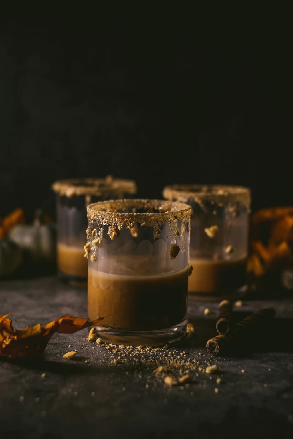Three glasses of pumpkin spice cocktails with sugar rims, dim lighting, and scattered crumbs, creating a moody atmosphere.