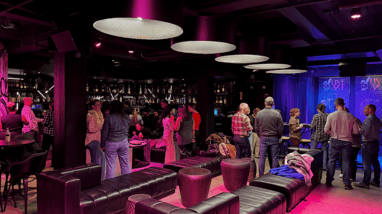 Guests mingle in a stylish lounge with purple lighting, leather seating, and a bar in the background at Live Axe.