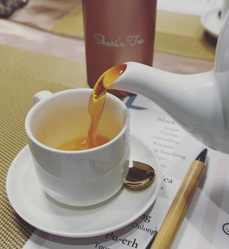 Close-up of tea being poured from a white teapot into a cup with a golden spoon, featuring a “Shari’s Tea” tumbler in the background and a tea menu listing premium blends on the table.
