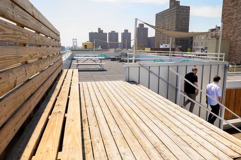 A bright daytime view of a rooftop deck in an urban setting with wooden bench seating in the foreground. The rooftop features picnic tables, blue accent chairs, and a tan sun shade. Two people in casual attire are walking down steps on the right. In the background, multiple residential high-rise apartment buildings are visible against a clear blue sky, with what appears to be the Williamsburg Bridge partially visible in the distance. The setting suggests a communal rooftop space in Brooklyn, New York.
