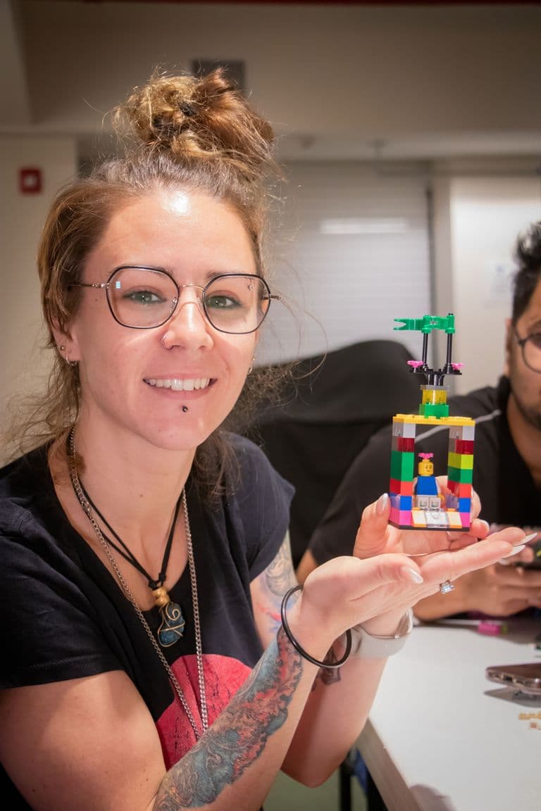 Smiling woman with glasses and tattoos holding up a colorful LEGO® creation featuring a mini-figure and green flag, seated at a table in an indoor setting