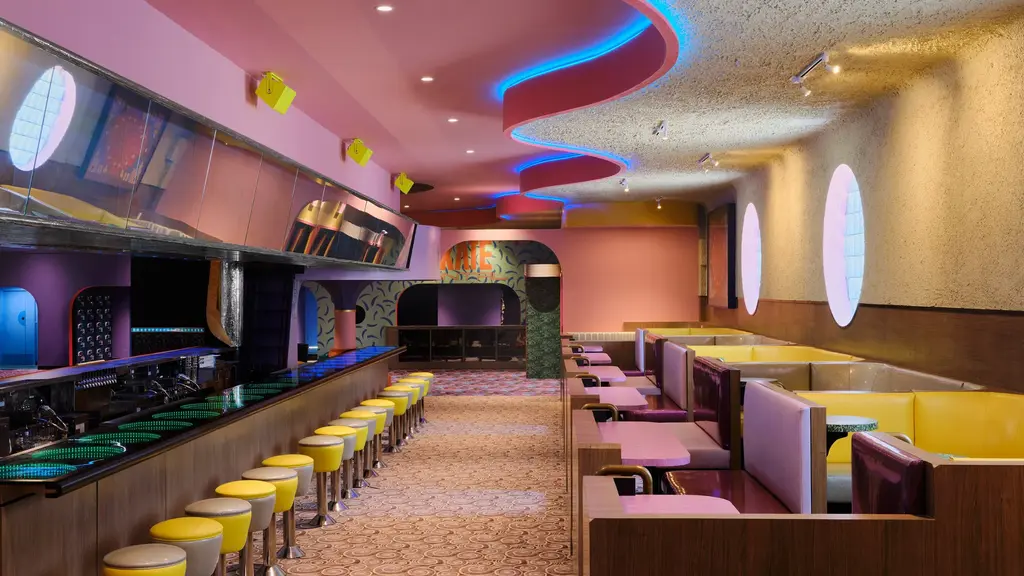 Retro-themed diner with a long bar, yellow stools, and colorful booths, featuring unique lighting and decor.